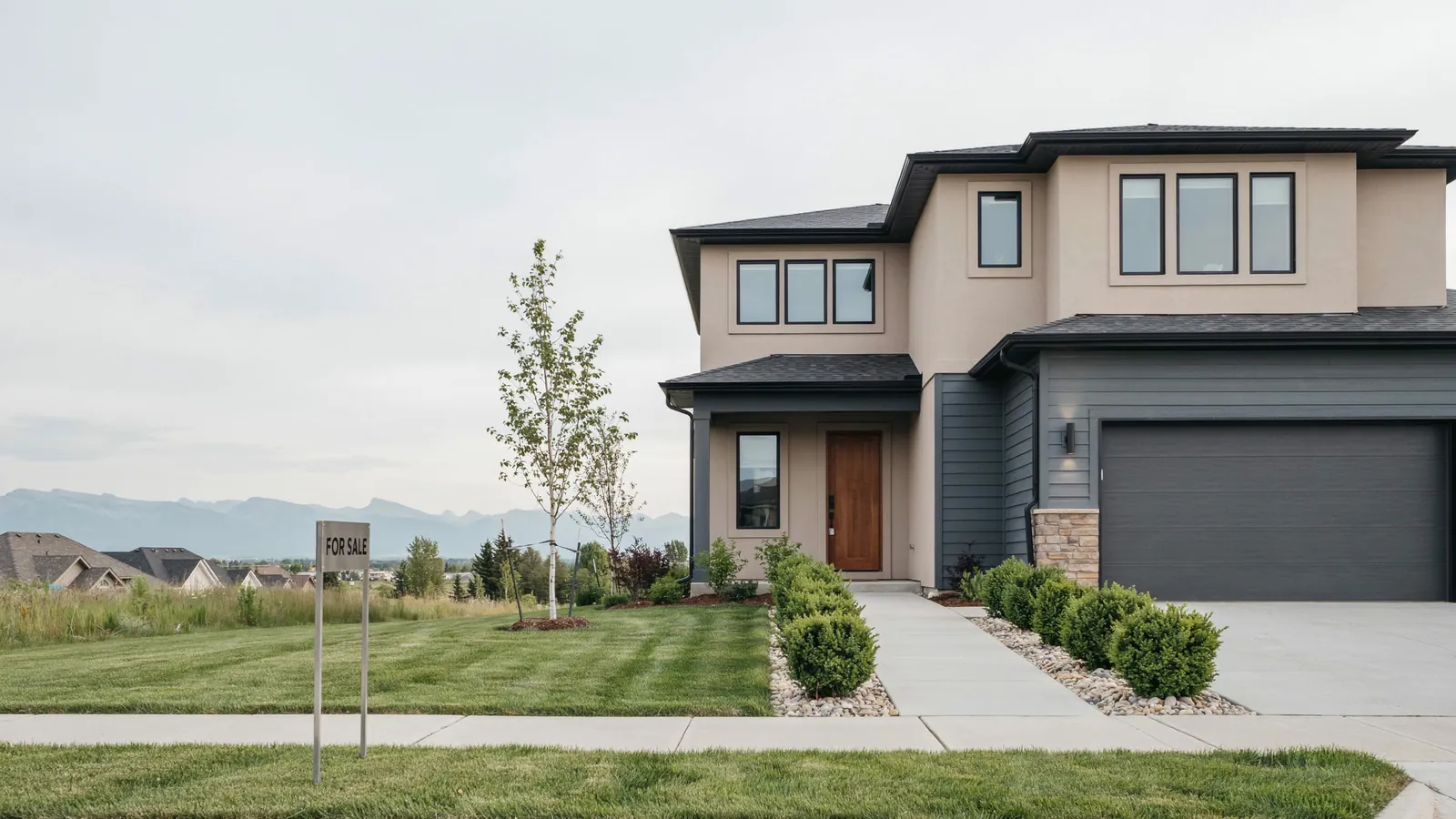 A modern two storey home with mountains behind it and a for sale sign on the lawn, the Edmonton foreclosure guide
